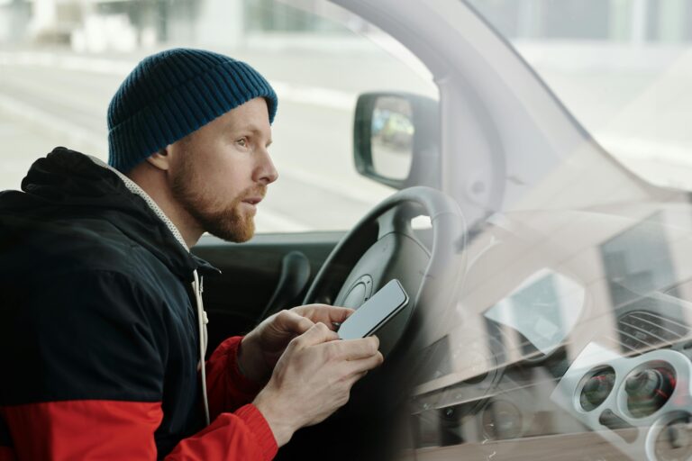 A man sitting in the driver’s seat of a car, holding a smartphone with a concerned expression, possibly after a traffic incident. The image highlights the importance of staying calm and using a phone to document an accident, contact emergency services, and communicate with insurance providers. Proper documentation can help ensure a smooth claims process and legal protection.