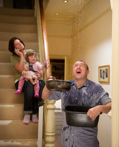 A family of three reacting to a roof leak in their home. Water drips from the ceiling into a bucket as the parents assess the damage, highlighting the need for home maintenance and proper insurance coverage.